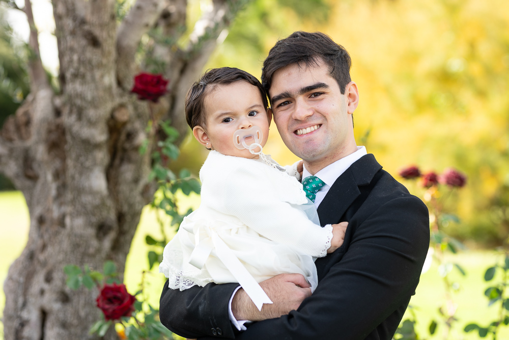 Primer plano de un joven padre con su hija en el dia de su Bautizo en un bonito jardin con un olivo y unos preciosos rosales